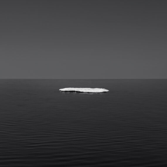Minimalist black and white photograph of a single small piece of melting sea ice floating on a vast, calm dark ocean under a clear gray sky
