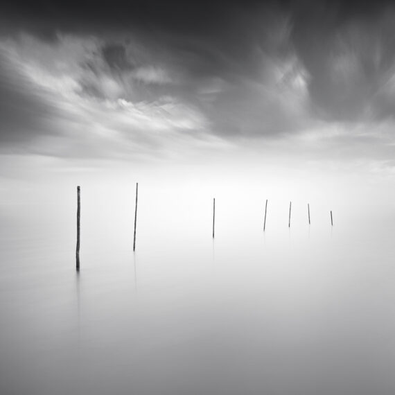 Minimalist black and white long exposure photograph of a diagonal row of thin wooden poles protruding from perfectly calm, misty water under a dramatic cloudy sky.