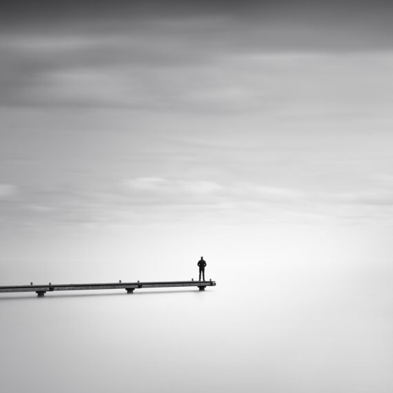 Minimalist black and white long exposure photograph of a solitary person standing at the edge of a long wooden pier, surrounded by a vast misty sky and calm water.