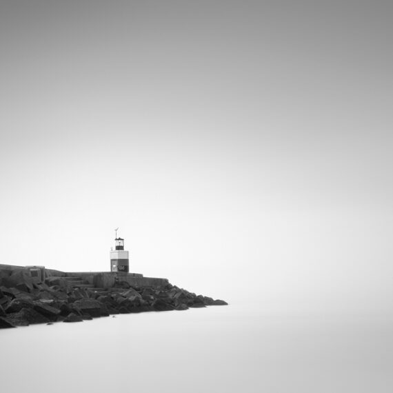 Minimalist black and white long exposure photograph of a small lighthouse at the end of a stone breakwater pier, surrounded by calm misty water.