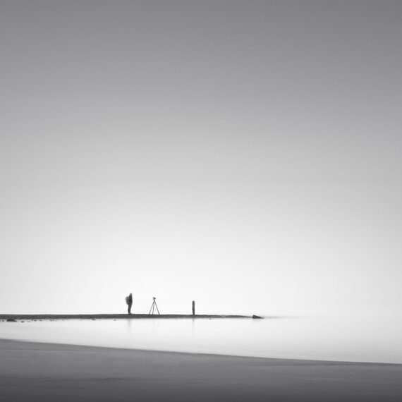 Minimalist black and white long exposure photograph of a photographer standing next to a camera on a tripod on a distant shoreline, with calm water and a vast misty sky.