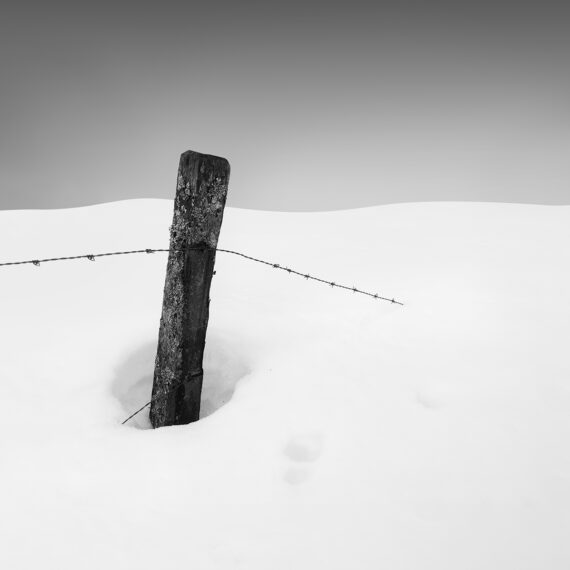 A wooden pole in the snow with barb wire disappearing in the snow